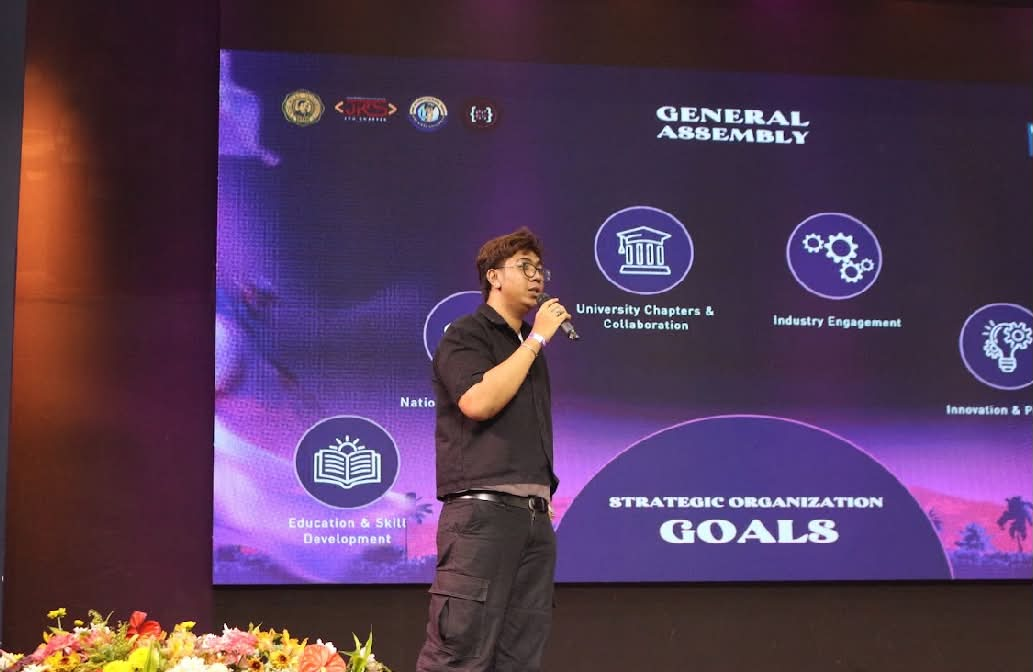 Miggy Rivera speaking into a microphone on stage at a general assembly.
